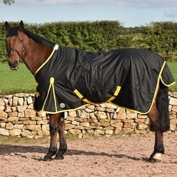 Black turnout rug with yellow surcingles on a bay horse by a stone wall